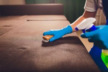 Cropped image. Cleaning concept. Male hand in light blue protective gloves cleaning sofa couch in the room.